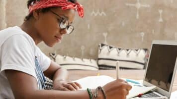 young African-American woman wearing glasses and head bandana, writing website content tips by hand in her notebook. Laptop is also visible in front of the woman.